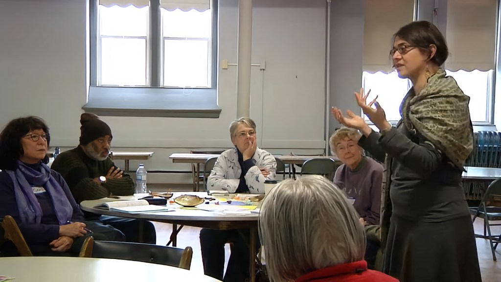 Photo of Naomi Cohn teaching while wearing a green shawl. She gestures with her hands while group of writers listen to her while seated at a round table.